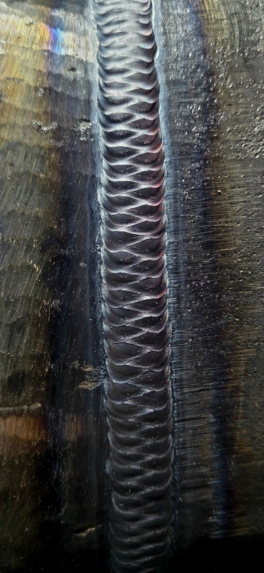 Szlifowana powierzchnia metalu z wyraźnym, idealnie równym śladem spoiny o regularnym wzorze falistym, widoczne tęczowe refleksy światła.