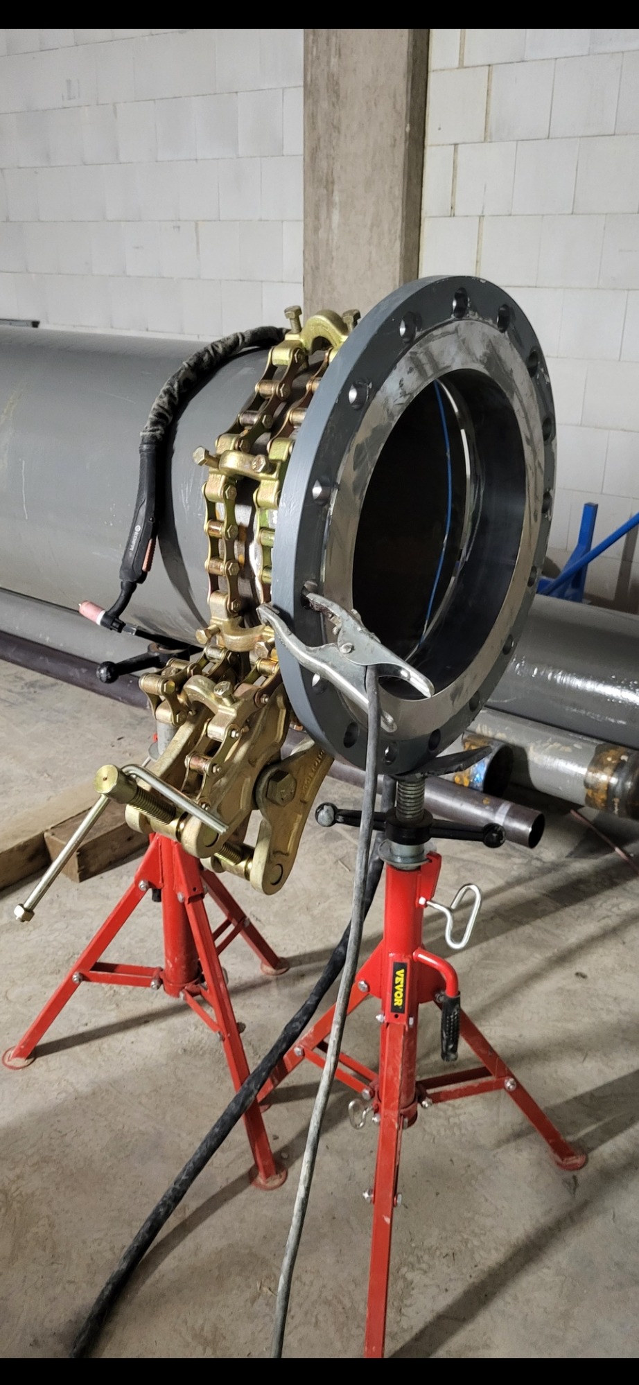 Przygotowanie do spawania rury z kołnierzem, wykorzystujące zacisk łańcuchowy na dwóch czerwonych stojakach, z widocznym uchwytem spawalniczym TIG i przewodami.