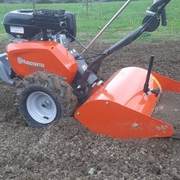 Dudziak. Wypożyczalnia sprzętu ogrodniczego i budownictwo - Pomarańczowy glebogryzarka Husqvarna TX 300 w trakcie pracy na polu, widoczne świeżo spulchnione grudy ziemi.