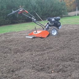 Dudziak. Wypożyczalnia sprzętu ogrodniczego i budownictwo - Pomarańczowy glebogryzarkę spalinową z czarnym silnikiem przygotowuje glebę na polu uprawnym, widok z boku, w tle drzewa i ogrodzenie.