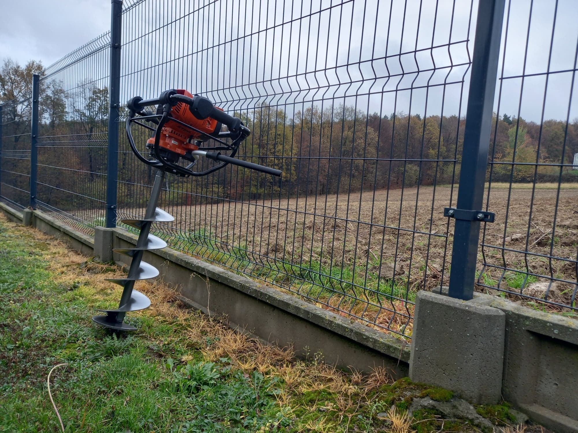 Wiertnica glebowa spalinowa Stihl BT 131, wypożyczona w Trzcianie, oparta o panel ogrodzeniowy z siatki.