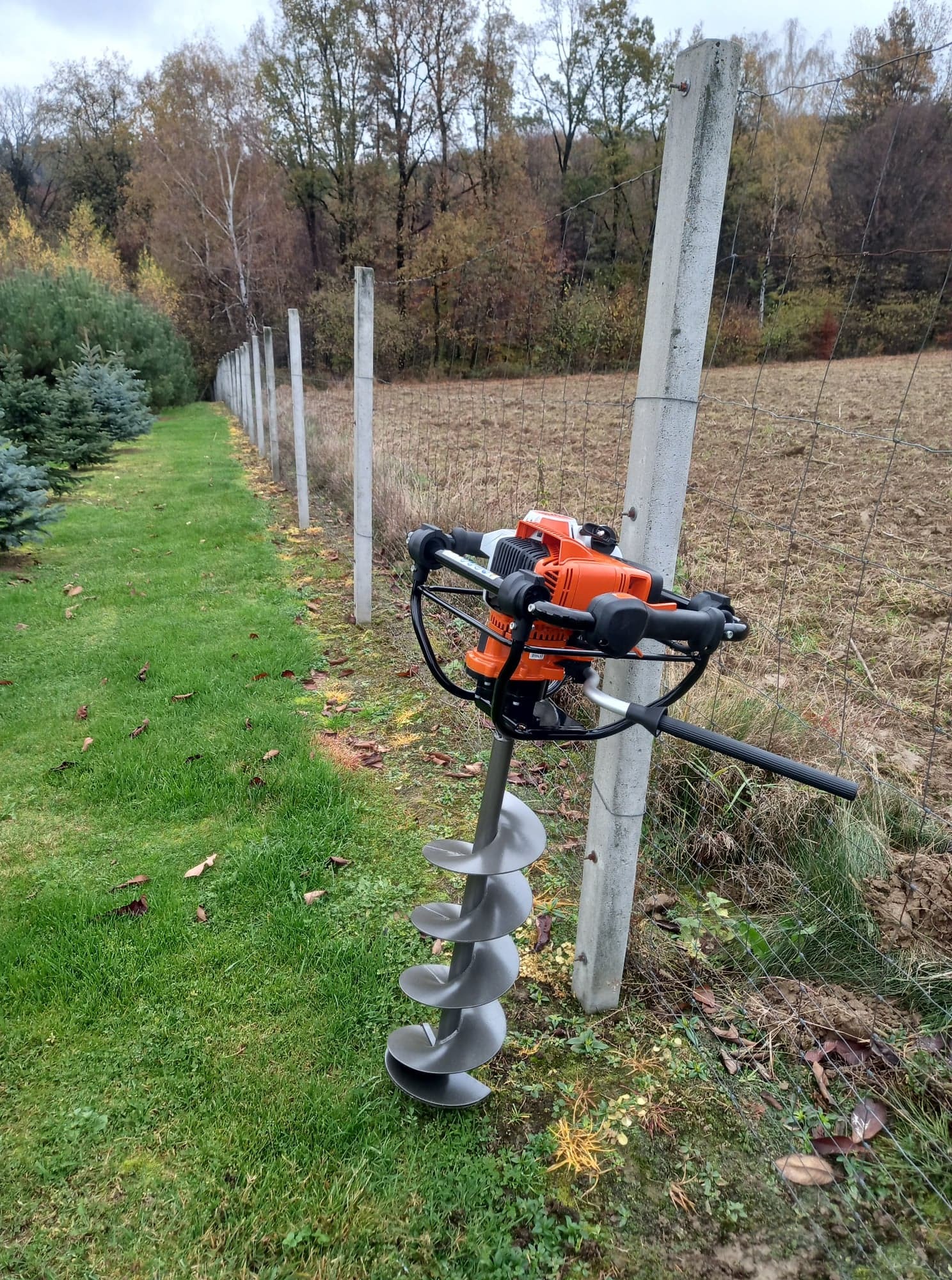 Wiertnica glebowa spalinowa oparta o betonowy słupek ogrodzenia z siatki, trawa i drzewa w tle.