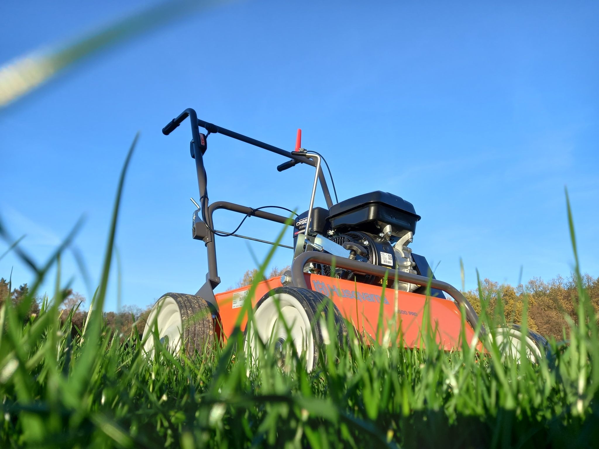 Wertykulator Husqvarna 500 PRO widziany z perspektywy trawy, z czarnym silnikiem i pomarańczową obudową na tle błękitnego nieba i drzew w oddali.
