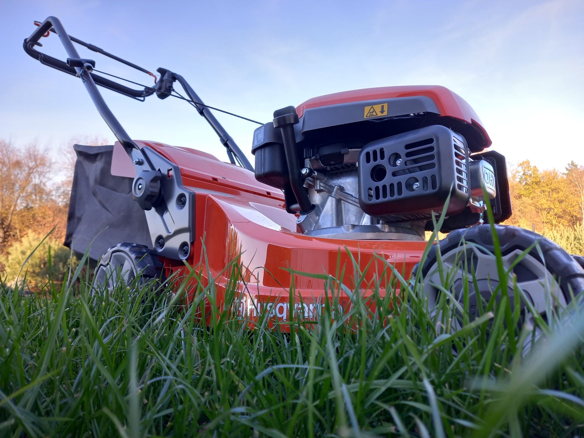 Pomarańczowa kosiarka spalinowa Husqvarna na trawie, widok z dołu, z widocznym silnikiem i koszem na trawę.