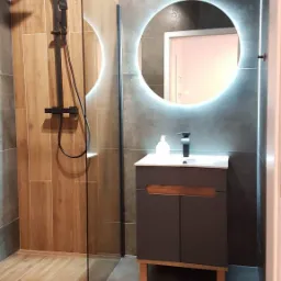 Nowoczesna łazienka z kabiną prysznicową typu walk-in, drewnopodobnymi płytkami ściennymi i podłogowymi, szarą ścianą z okrągłym lustrem LED nad szafką z umywalką.