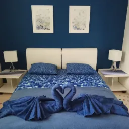 Niebieska sypialnia z łóżkiem, na którym ułożono dwa łabędzie z ręczników, białe szafki nocne z lampkami i dwa obrazy na ścianie.