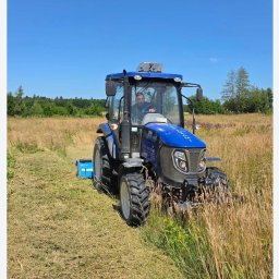 Pod Brzozami Jakub Grajek - Niebieski traktor LOVOL M54 z operatorem na polu w słoneczny dzień, częściowo skoszone trawy. Widoczne drzewa w tle i fragment nieba.