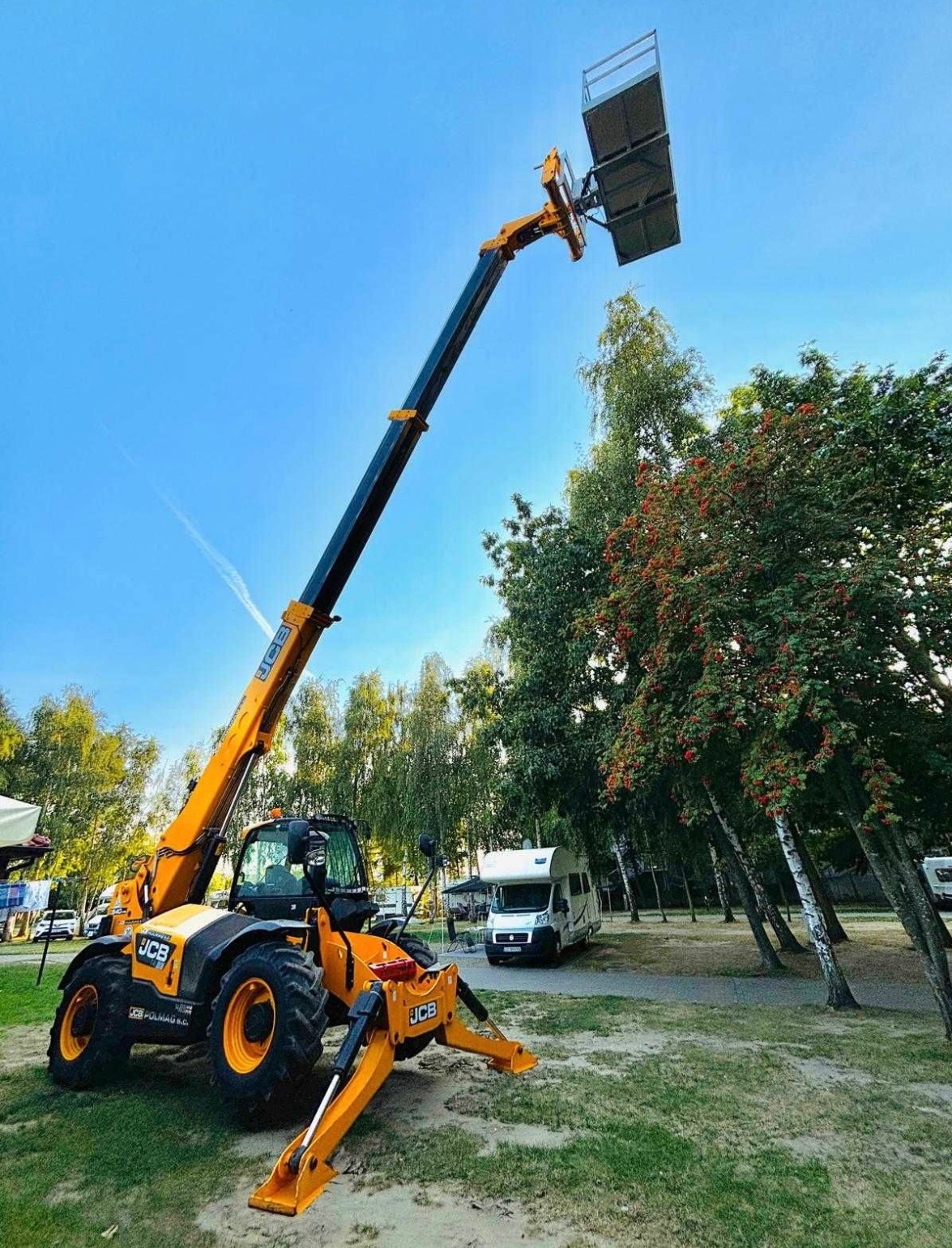 Żółta podnośnik koszowy JCB z wysięgnikiem na tle błękitnego nieba i zieleni drzew, z platformą roboczą na końcu ramienia. W tle widoczny kamper.