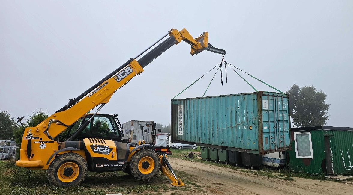 Żółta ładowarka teleskopowa JCB podnosi turkusowy kontener transportowy na zewnątrz, w tle widoczne inne kontenery i częściowo zamglone niebo.