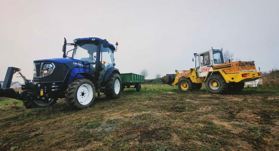 Niebieski traktor z przyczepą i żółta ładowarka na polu w pochmurny dzień. Maszyny gotowe do pracy.