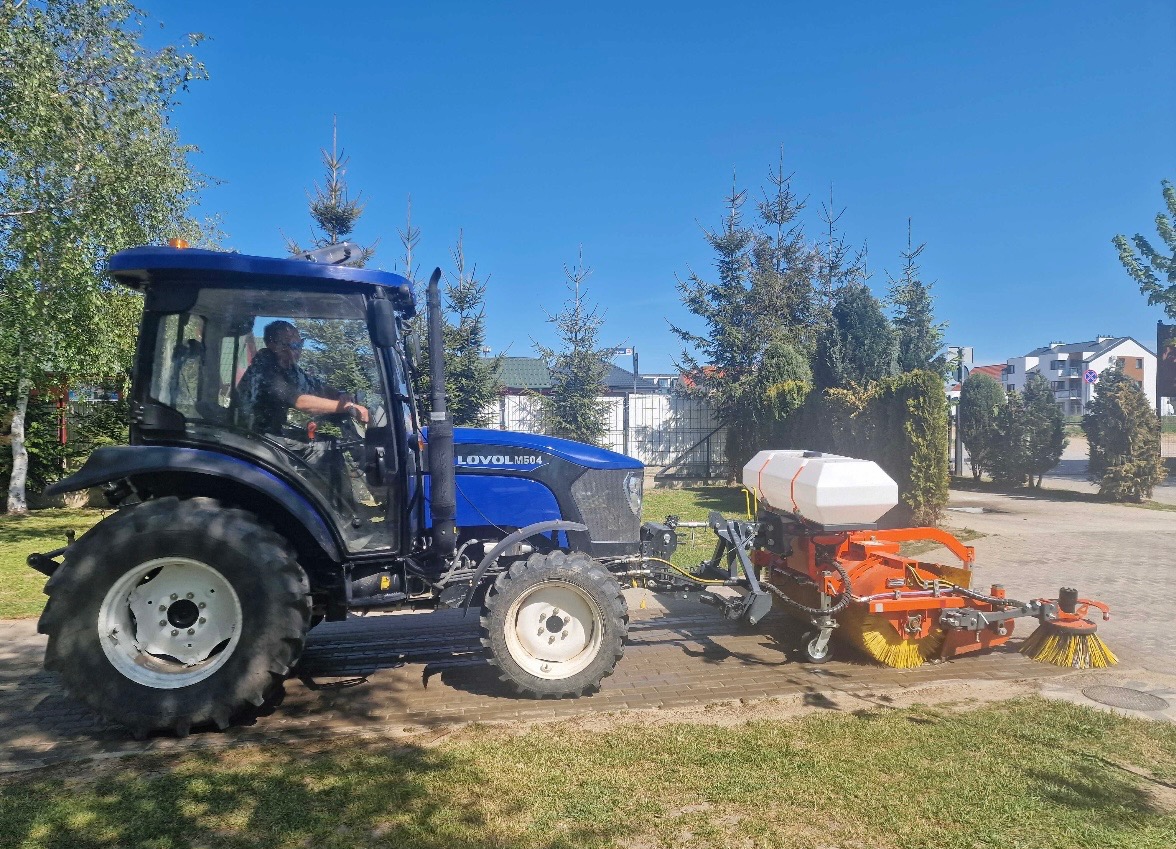 Niebieski traktor Lovol M504 z pomarańczową szczotką czyszczącą, obsługiwany przez operatora, na tle kostki brukowej i zieleni w słoneczny dzień.