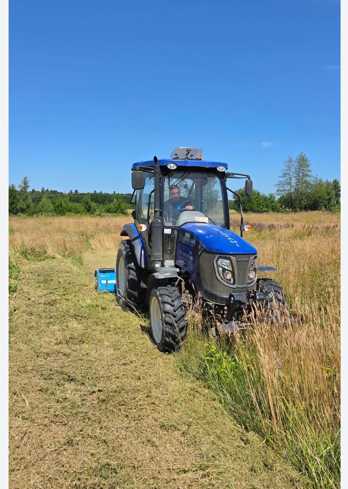Niebieski traktor LOVOL M54 z operatorem na polu w słoneczny dzień, częściowo skoszone trawy. Widoczne drzewa w tle i fragment nieba.