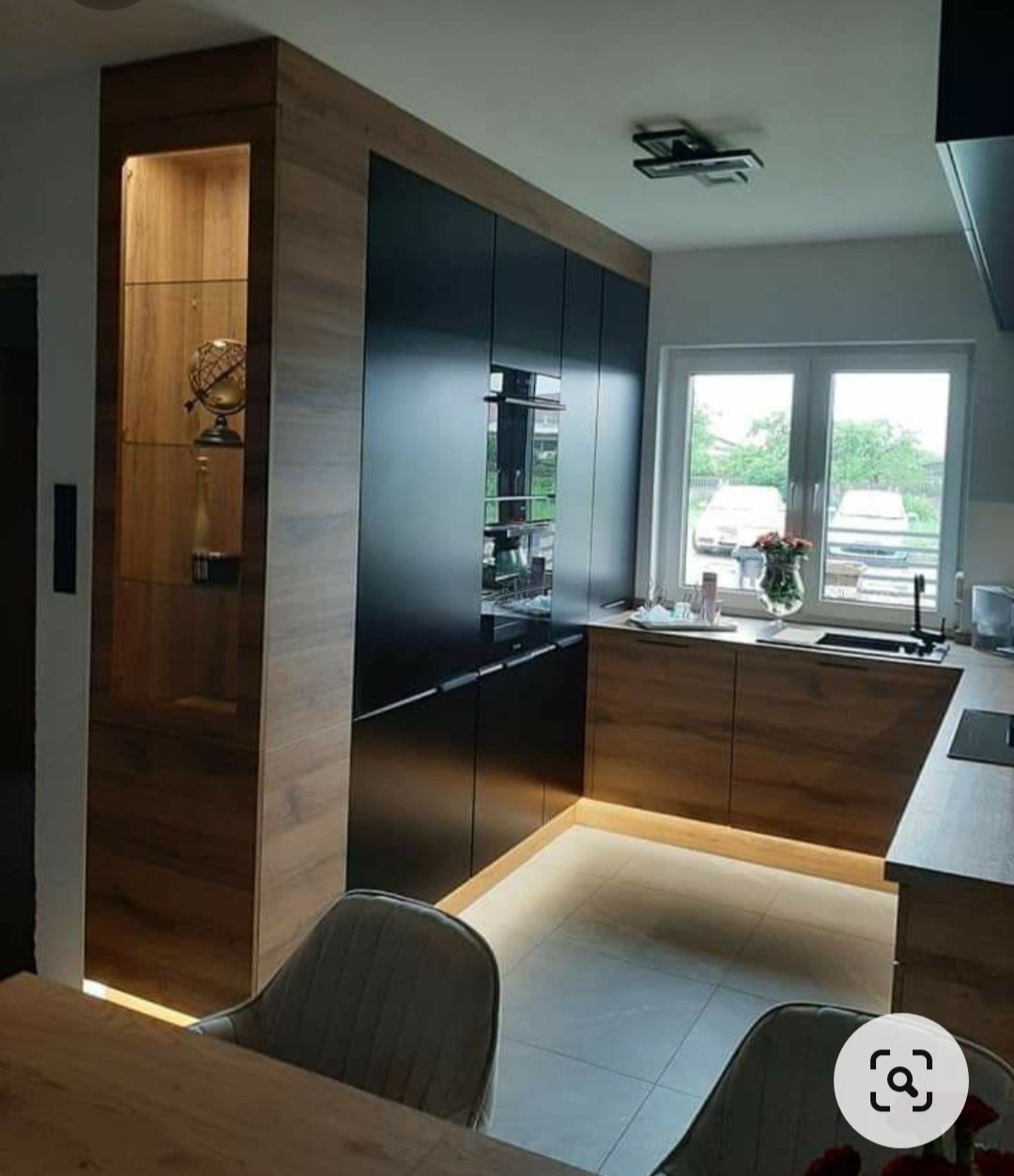 Nowoczesna kuchnia: czarne, matowe fronty szafek zestawione z drewnopodobnymi panelami, podświetlenie LED na podłodze, szklana witryna z dekoracjami, widok na okno z kwiatami.