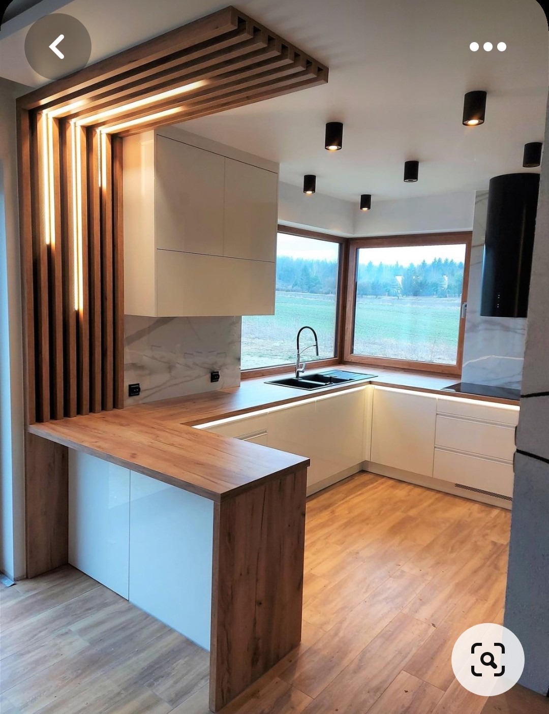 Nowoczesna kuchnia z białymi szafkami, drewnianym blatem i dekoracyjnymi elementami oświetleniowymi, z widokiem na zewnątrz przez duże okno.