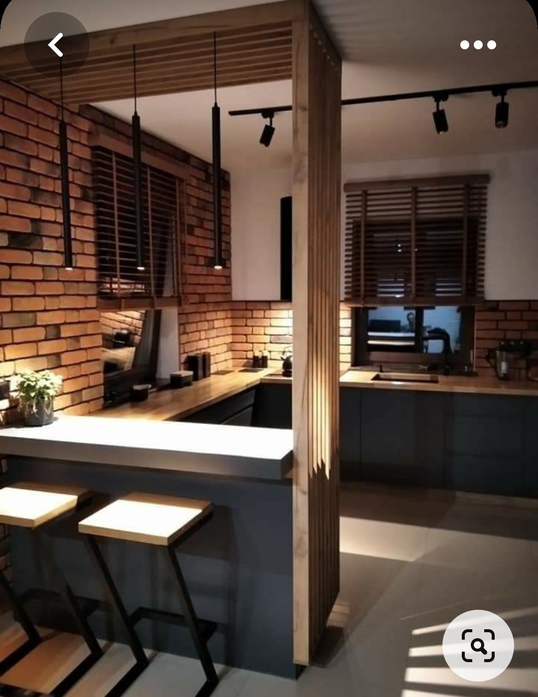 Nowoczesna kuchnia z ceglaną ścianą, drewnianym blatem i barkiem z dwoma hokerami, oświetlona wiszącymi lampami i reflektorami, oddzielona drewnianą ażurową ścianką.