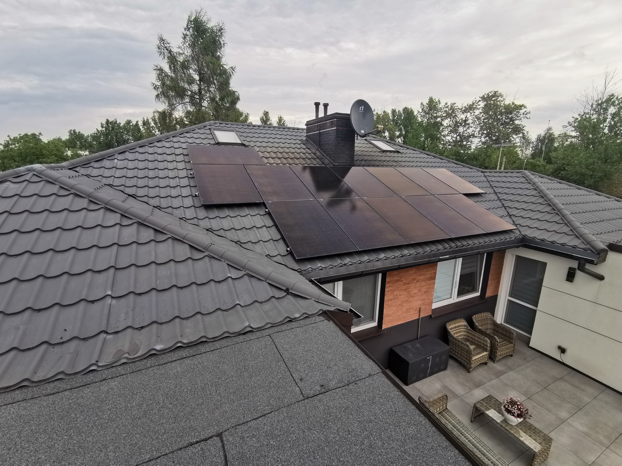 Czarny dach z dachówki z zainstalowanymi panelami słonecznymi, komin z anteną satelitarną, widok z góry na patio z meblami ogrodowymi i drzewa w tle.