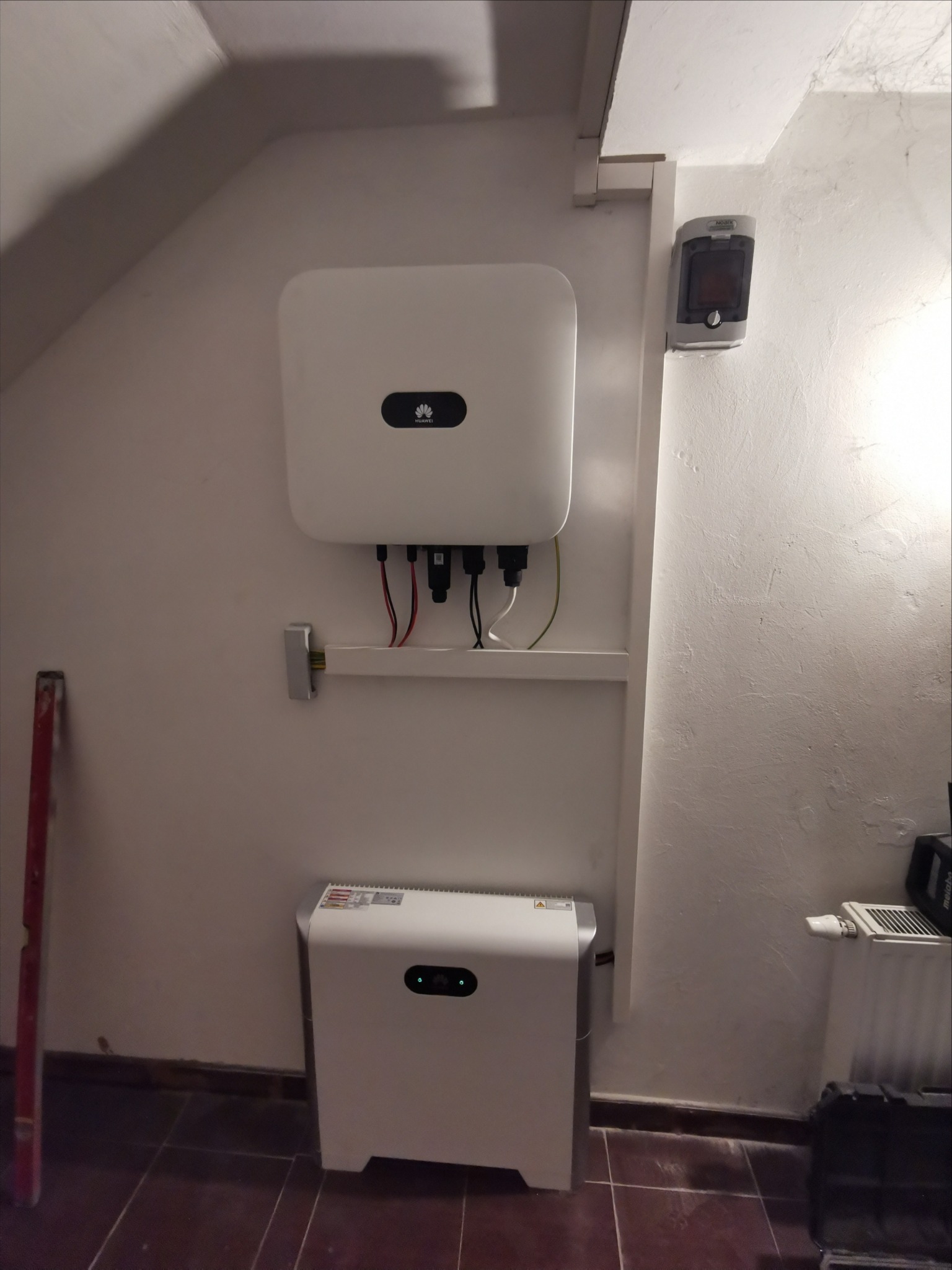 Zamontowany biały inwerter fotowoltaiczny Huawei z podłączonymi kablami, umieszczony nad białą obudową prawdopodobnie magazynu energii, w pomieszczeniu z białymi ścianami i brązową podłogą z płytek...