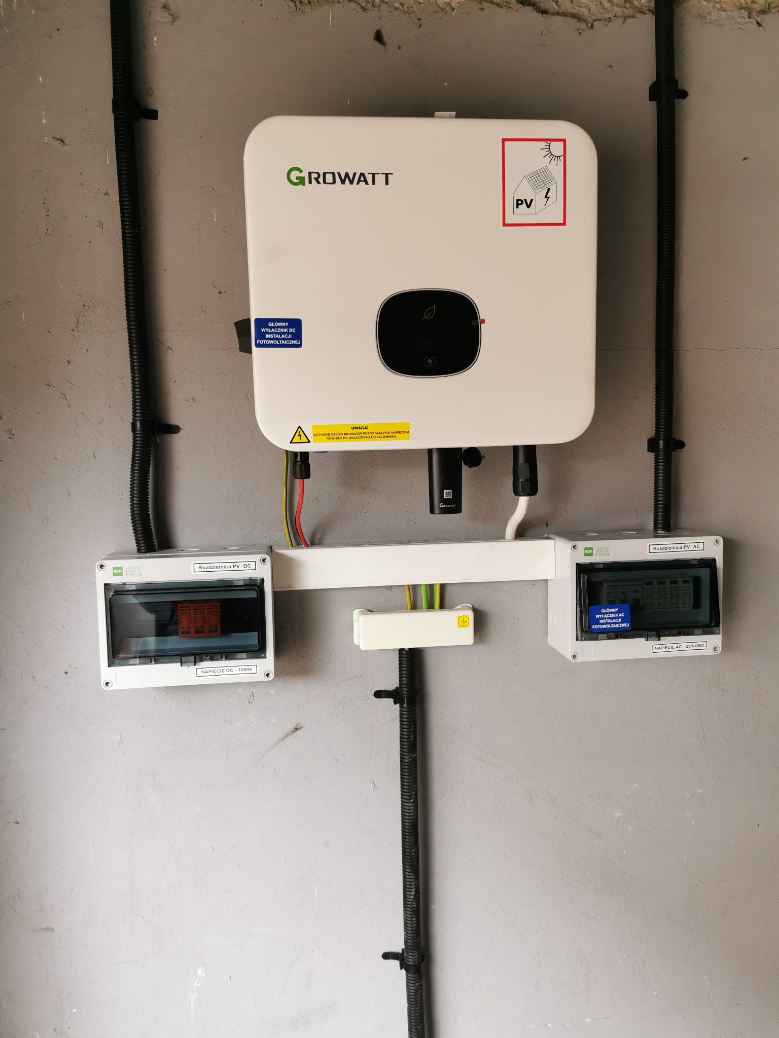 Biały falownik Growatt zamontowany na szarej ścianie, z rozdzielnicami PV-DC i PV-AC po bokach, połączony czarnymi rurkami instalacyjnymi.