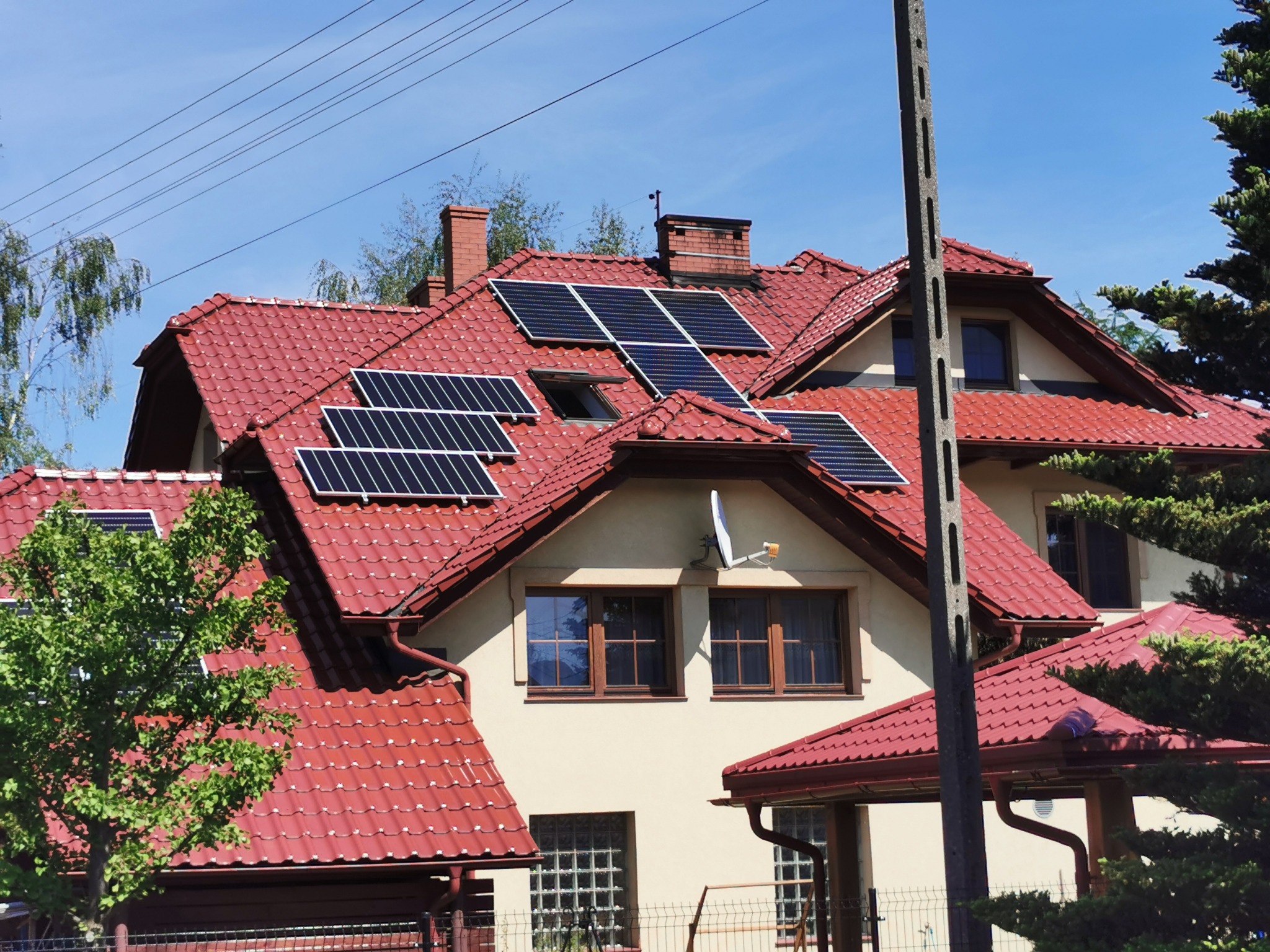 Dom jednorodzinny z czerwoną dachówką, na której zamontowane są panele słoneczne; widoczna antena satelitarna, drzewa i słup energetyczny.