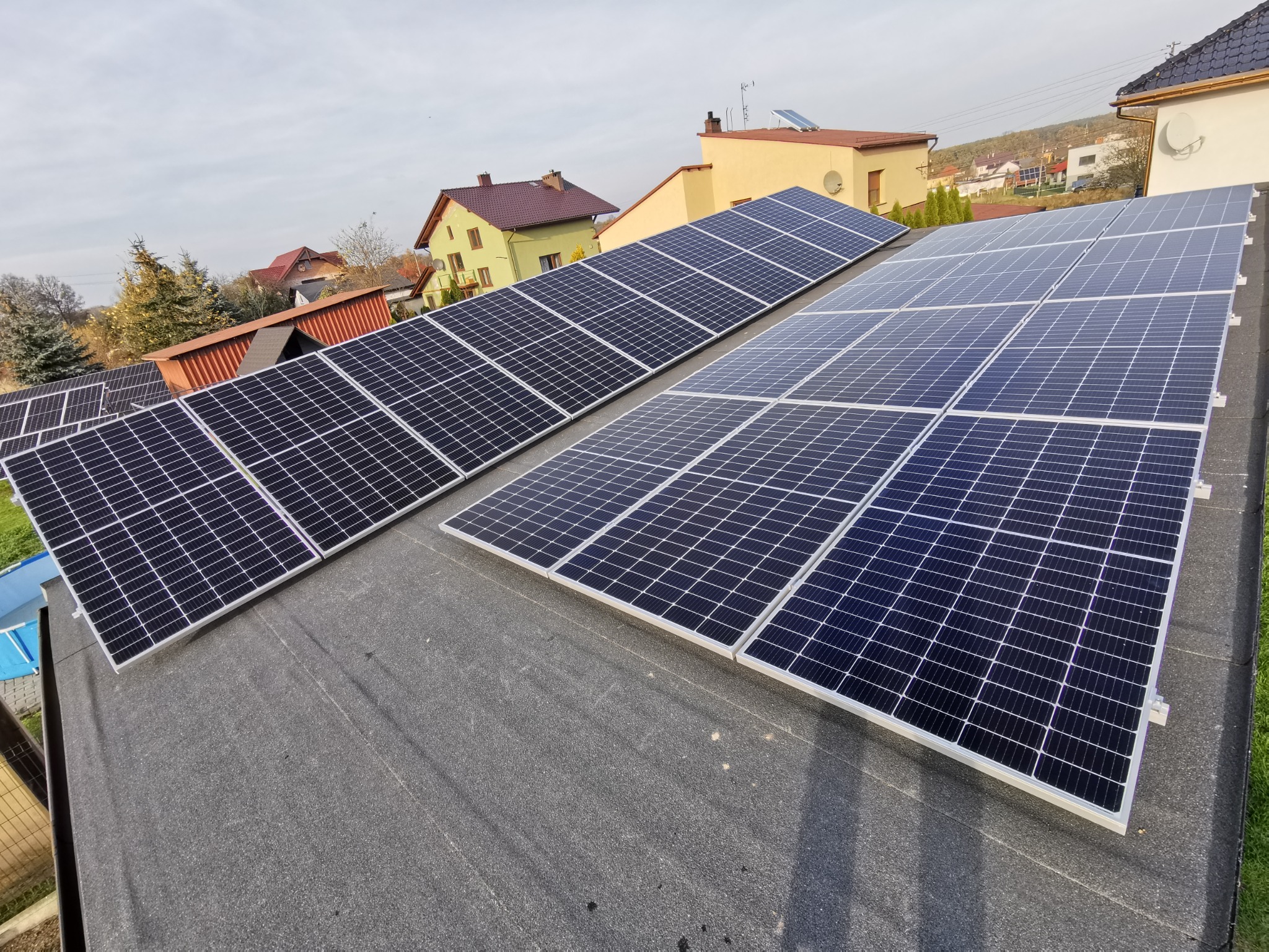 Dwa rzędy paneli słonecznych na płaskim, bitumicznym dachu, widok z góry, w tle zabudowa jednorodzinna i pochmurne niebo.