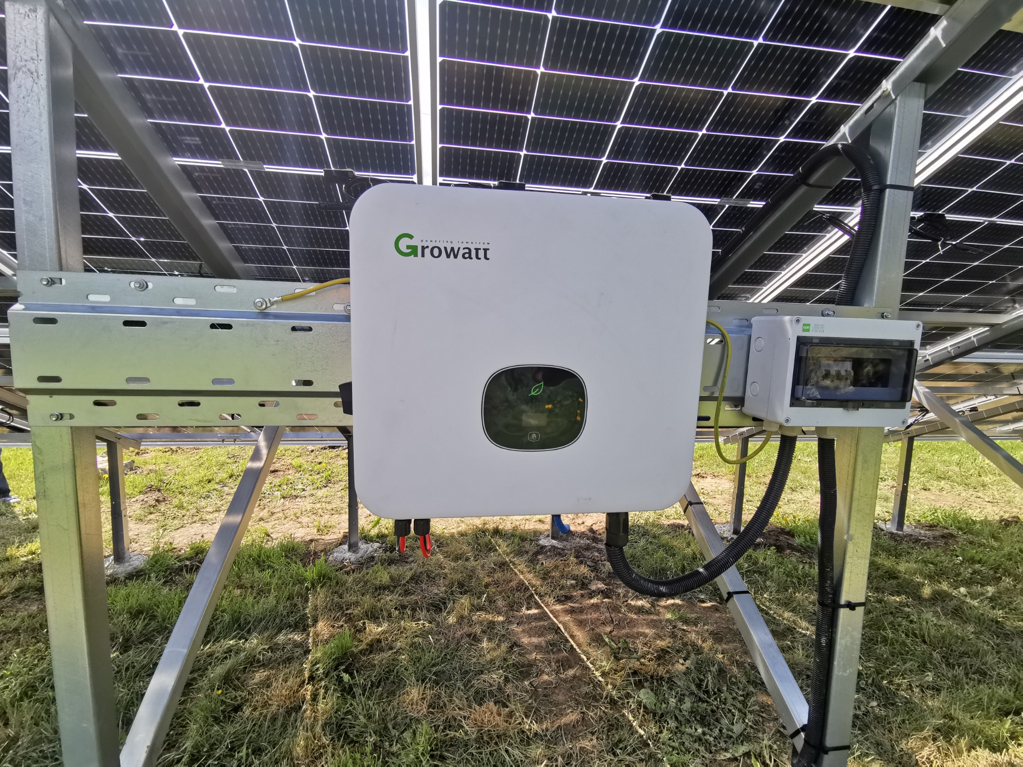 Biały falownik Growatt zamontowany pod panelami fotowoltaicznymi na metalowej konstrukcji, widoczne okablowanie i skrzynka zabezpieczeń, trawa w tle.