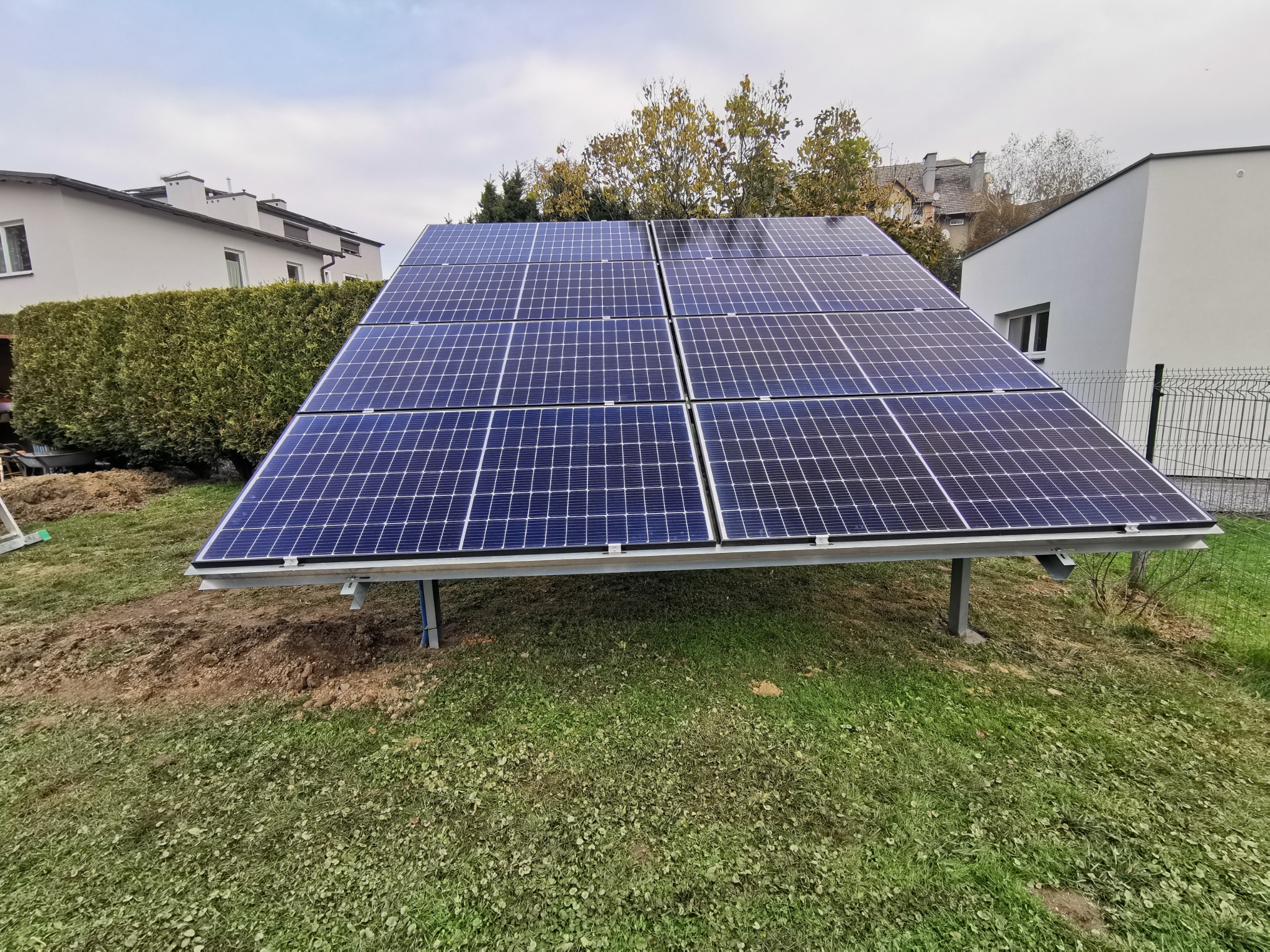 Instalacja paneli słonecznych na trawniku w Lubomi, widok na domy w tle, świeżo wykopana ziemia wokół konstrukcji wsporczej.