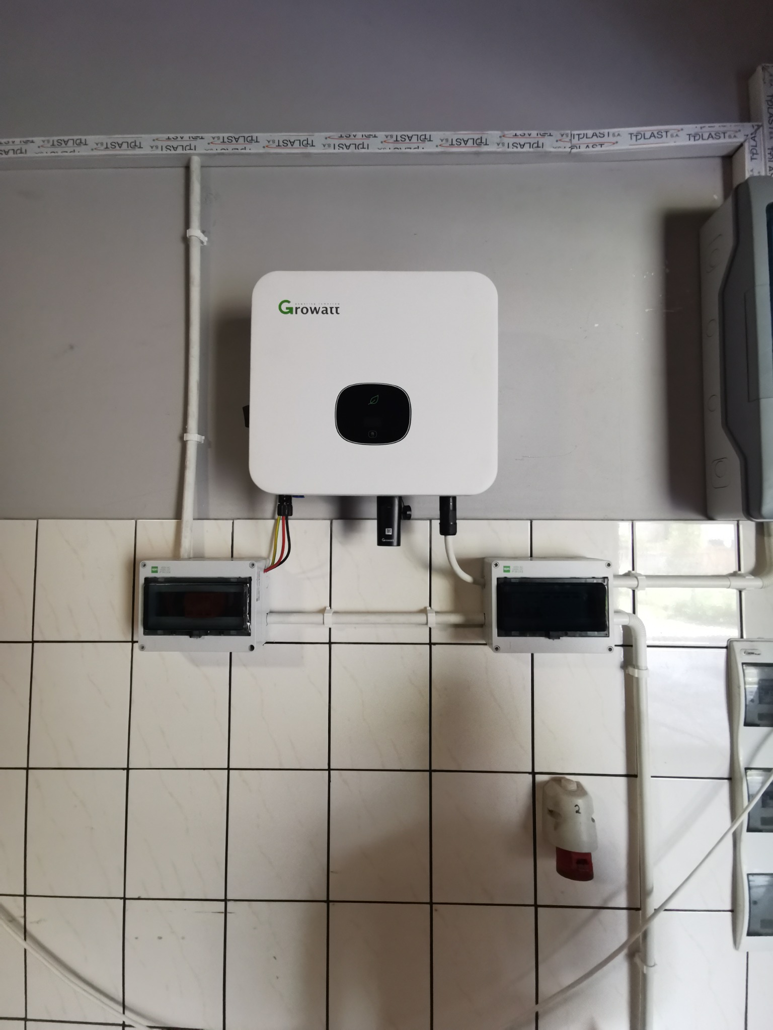 Zamontowany biały falownik Growatt na ścianie z białych płytek, widoczne kable, rurki instalacyjne i dwie skrzynki elektryczne, instalacja wewnątrz budynku.