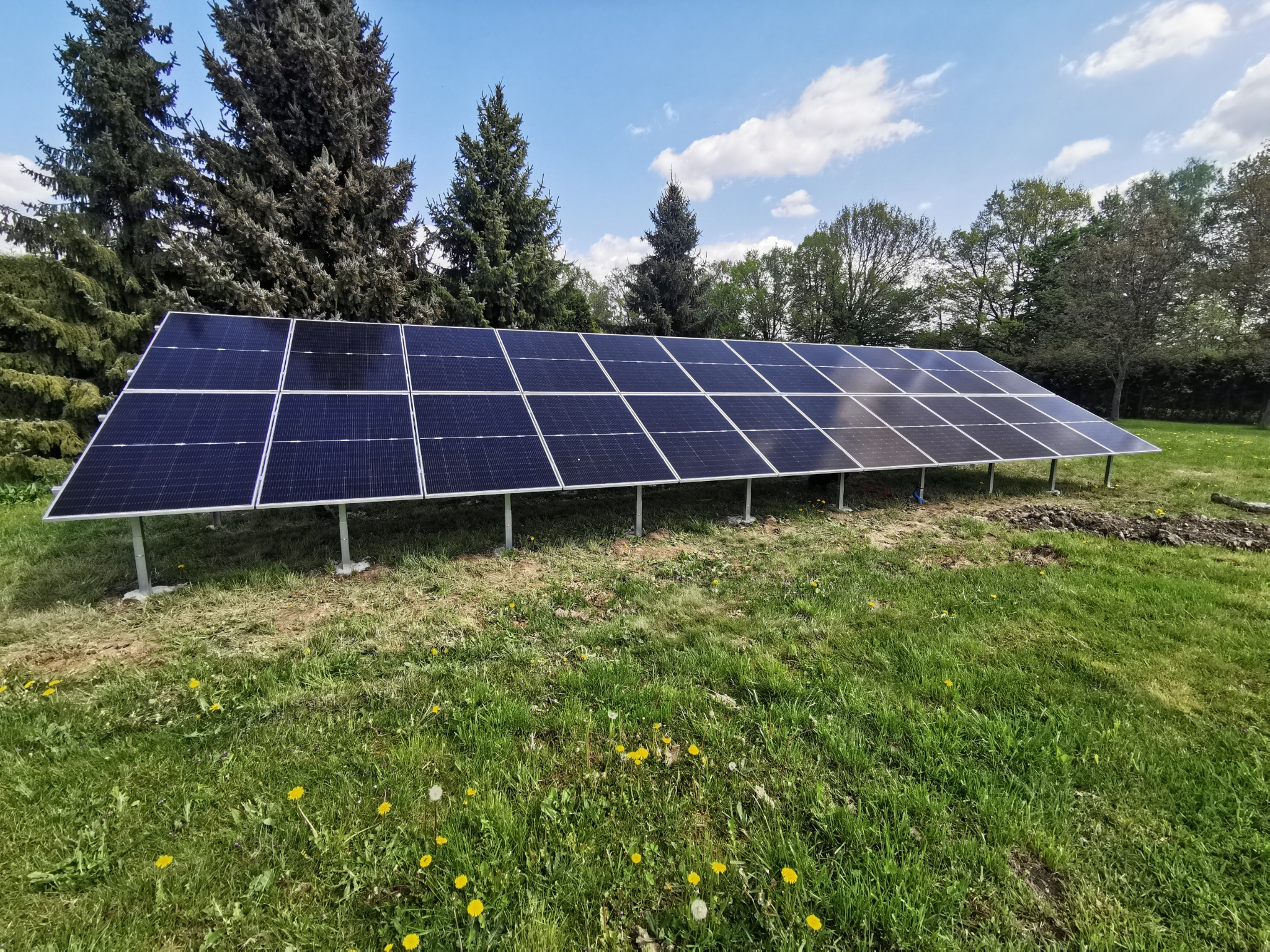 Rząd paneli słonecznych ustawionych na metalowych wspornikach na trawiastym terenie z drzewami w tle i błękitnym niebem z chmurami.