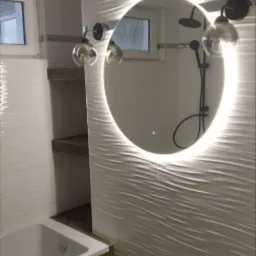 Nowoczesna łazienka z owalnym lustrem podświetlanym LED, białe płytki 3D na ścianach, bateria prysznicowa w kolorze czarnym widoczna w odbiciu lustra, drewnopodobna obudowa wanny i instalacja wodna...