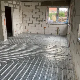 Białe rury ogrzewania podłogowego ułożone na czarnej siatce w surowym wnętrzu budynku z białych bloczków, widoczne okno i instalacje elektryczne w ścianach.