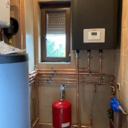 Czarny piec grzewczy marki Stiebel Eltron z wyświetlaczem, podłączony miedzianymi rurami do instalacji, z czerwonym zbiornikiem wyrównawczym i filtrami wody w pomieszczeniu wyłożonym beżowymi...