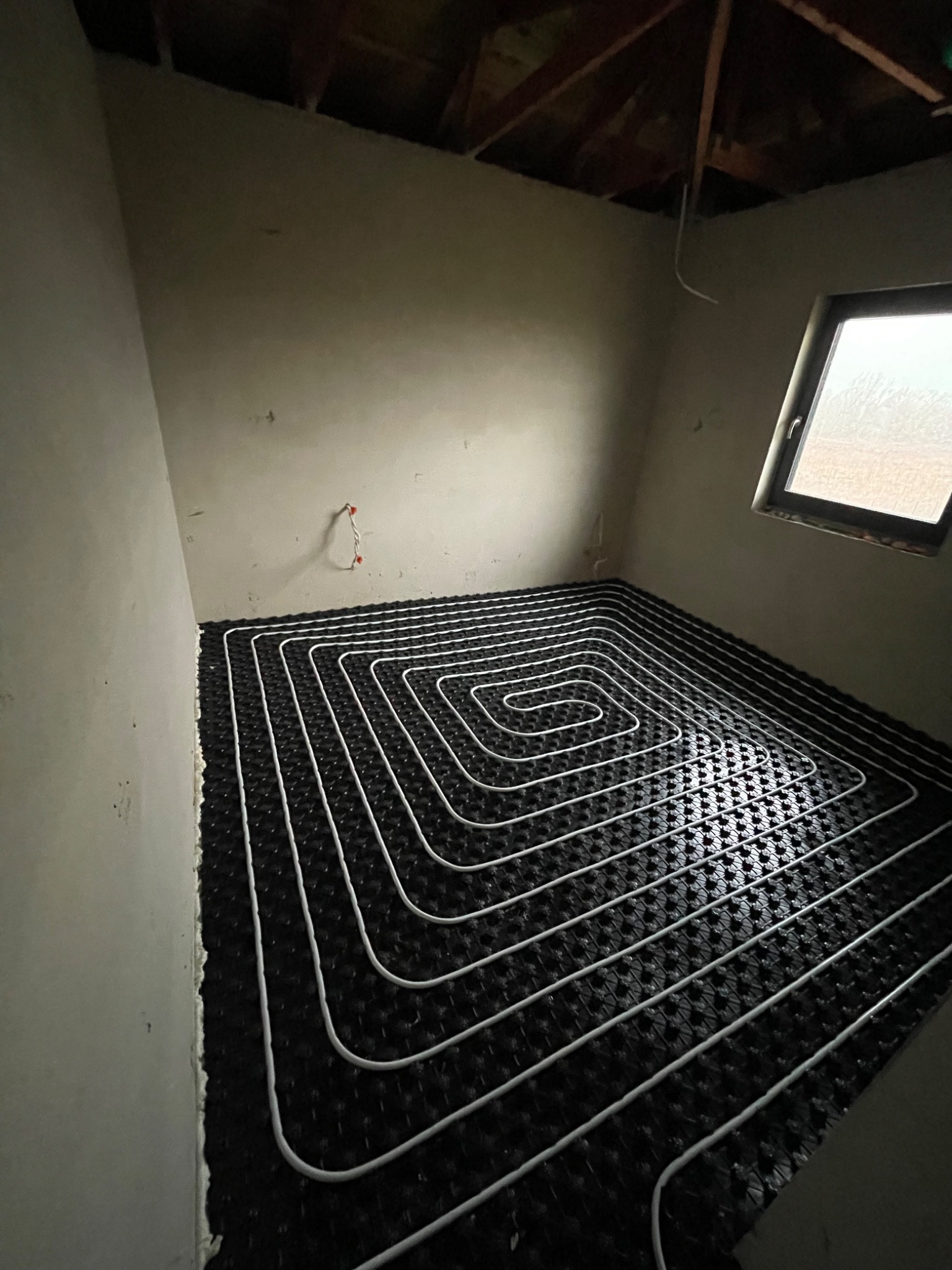 Białe rurki ogrzewania podłogowego ułożone spiralnie na czarnej macie w surowym pomieszczeniu z widocznym oknem i fragmentem drewnianej konstrukcji stropu.