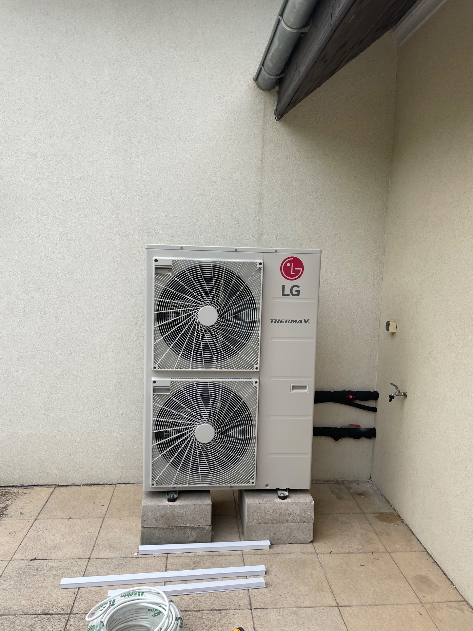 Zewnętrzna jednostka pompy ciepła LG Therma V, umieszczona na betonowych bloczkach, z widocznymi rurami w izolacji i elementami instalacji elektrycznej na tle elewacji budynku.