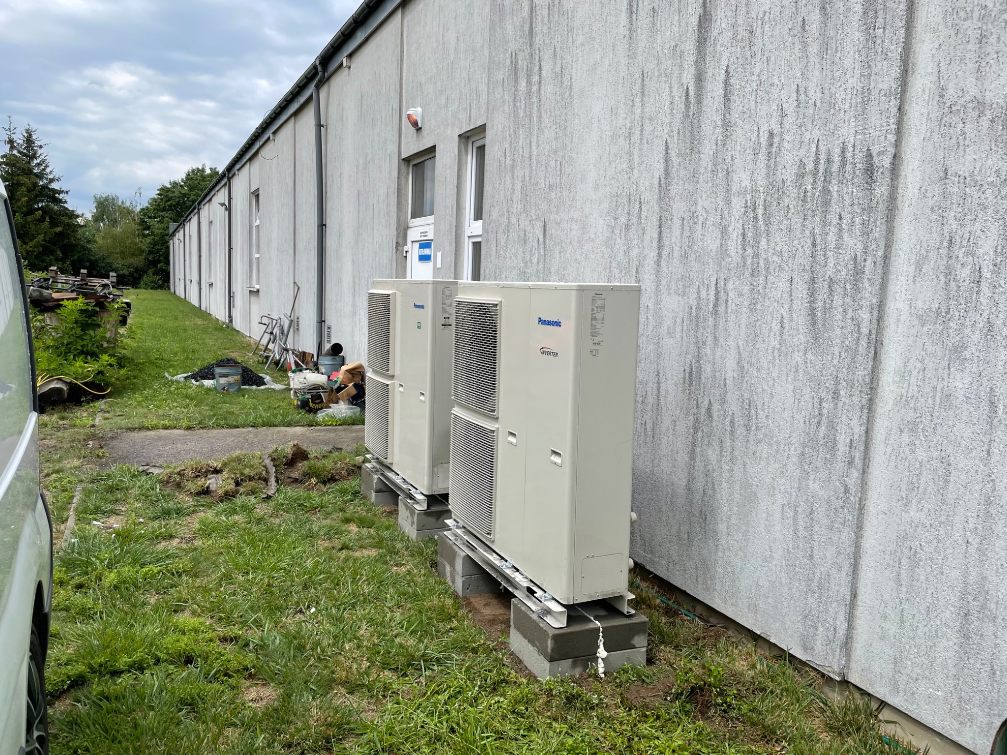 Dwa zewnętrzne moduły klimatyzacji Panasonic Inverter ustawione na betonowych blokach przy szarej ścianie budynku, widoczne ślady po montażu w trawie.