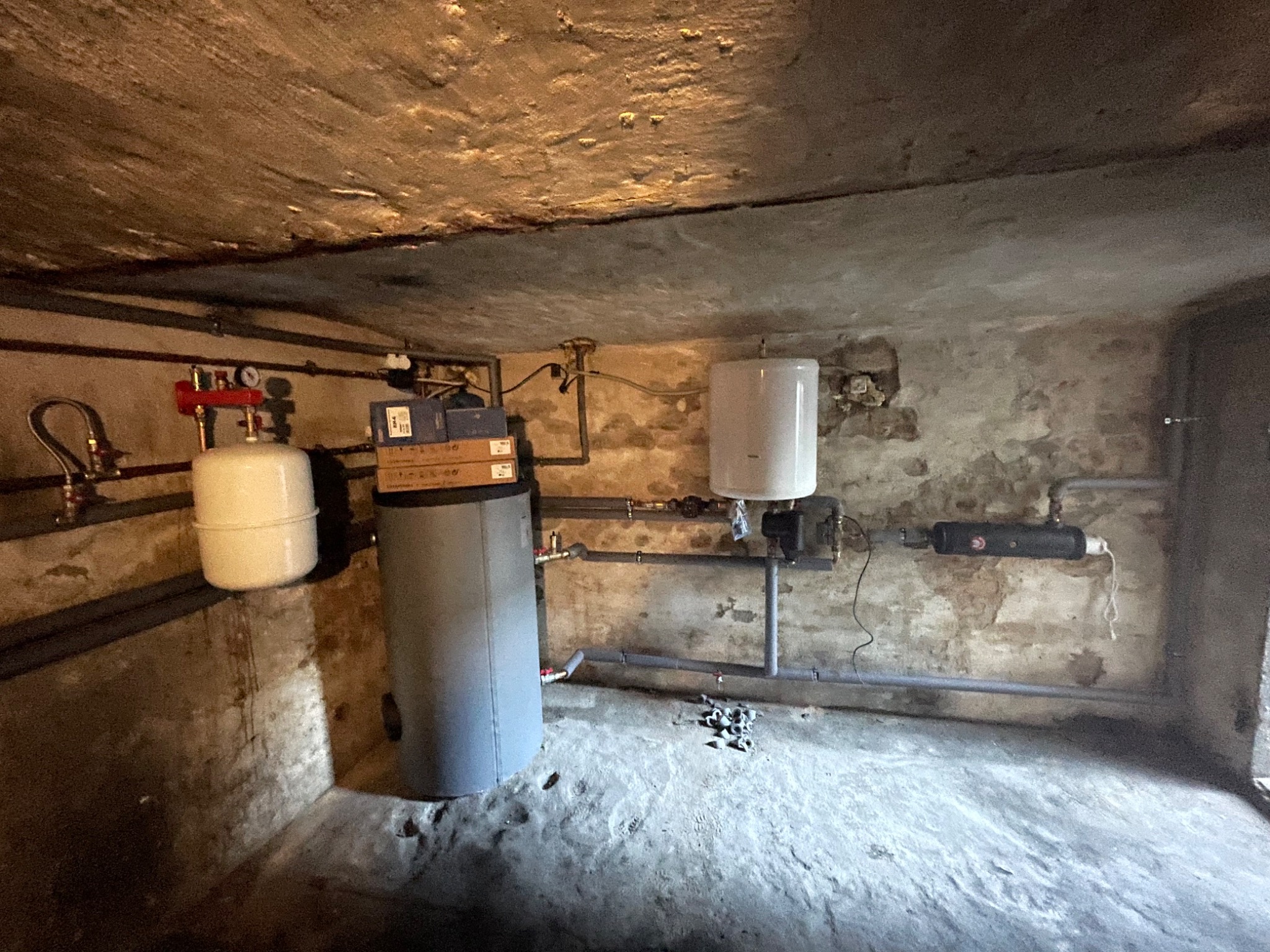 Instalacja grzewcza w piwnicy z widocznym zbiornikiem wyrównawczym, pompą obiegową, filtrem oraz rurami z tworzywa sztucznego. Na podłodze leżą złączki.