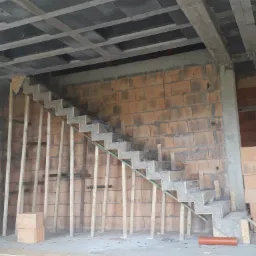 Niedokończone betonowe schody ze szkieletem z desek w surowym stanie, widok na ceglaną ścianę i betonowy strop.