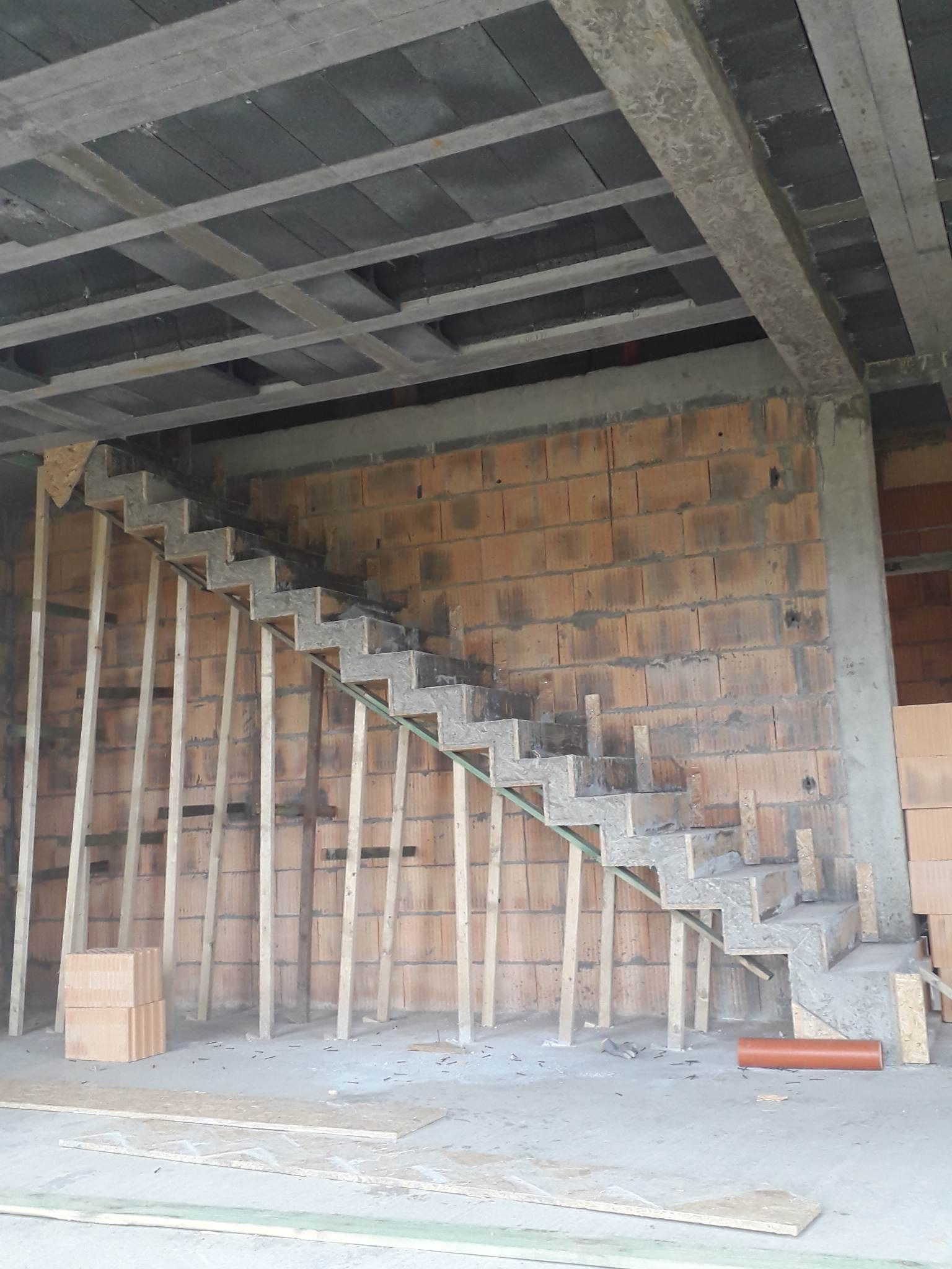 Niedokończone betonowe schody ze szkieletem z desek w surowym stanie, widok na ceglaną ścianę i betonowy strop.