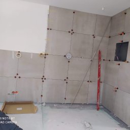 Tomili - Ściana w trakcie układania szarych płytek, z widocznymi krzyżykami dystansowymi i poziomnicą, fragment instalacji elektrycznej w ścianie, w tle widoczna płyta karton gips.
