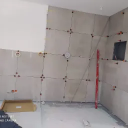 Ściana w trakcie układania szarych płytek, z widocznymi krzyżykami dystansowymi i poziomnicą, fragment instalacji elektrycznej w ścianie, w tle widoczna płyta karton gips.