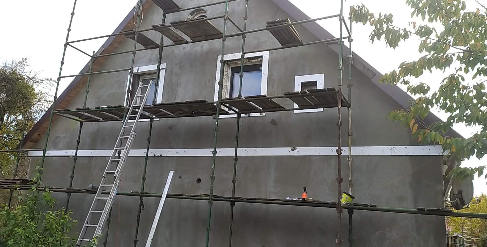 Dom w trakcie ocieplania, widoczne rusztowania, świeżo nałożony tynk w kolorze szarym, białe obramowania okien i biały pas na elewacji, drabina oparta o ścianę.