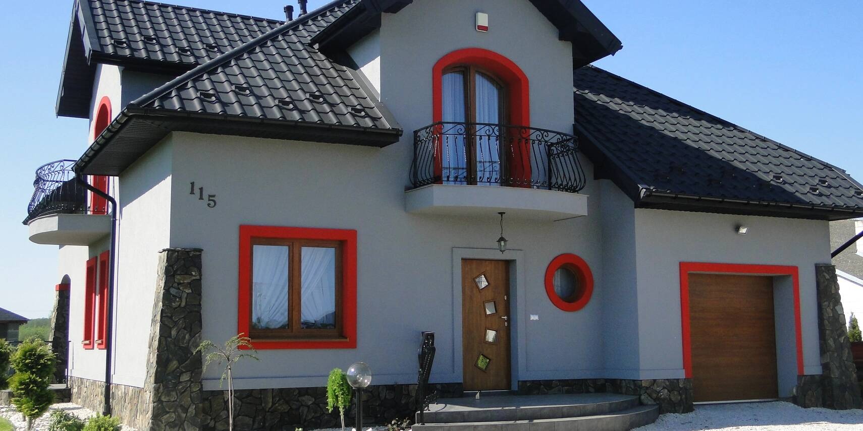 Szary dom jednorodzinny z czarnym dachem, czerwonymi obramowaniami okien i elementami dekoracyjnymi z kamienia na elewacji, numer domu 115.