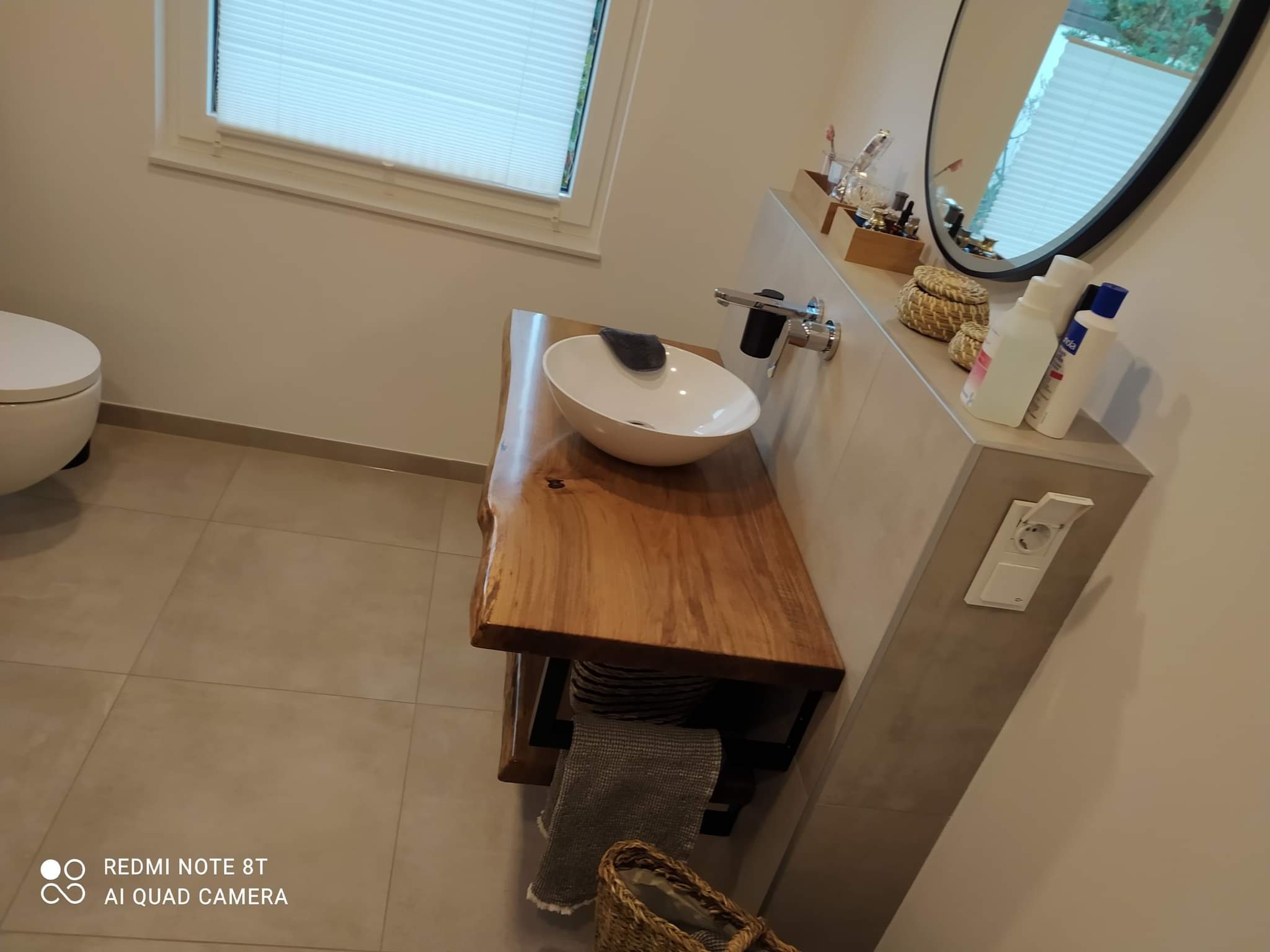 Nowoczesna łazienka z umywalką nablatową na blacie z surowego drewna, chromowaną baterią i okrągłym lustrem na ścianie wyłożonej płytkami w neutralnym odcieniu.