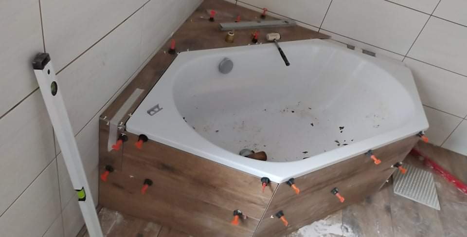 Montaż obudowy wanny z płytek imitujących drewno, z użyciem poziomicy i systemu poziomowania płytek; widoczne narzędzia i przybory.