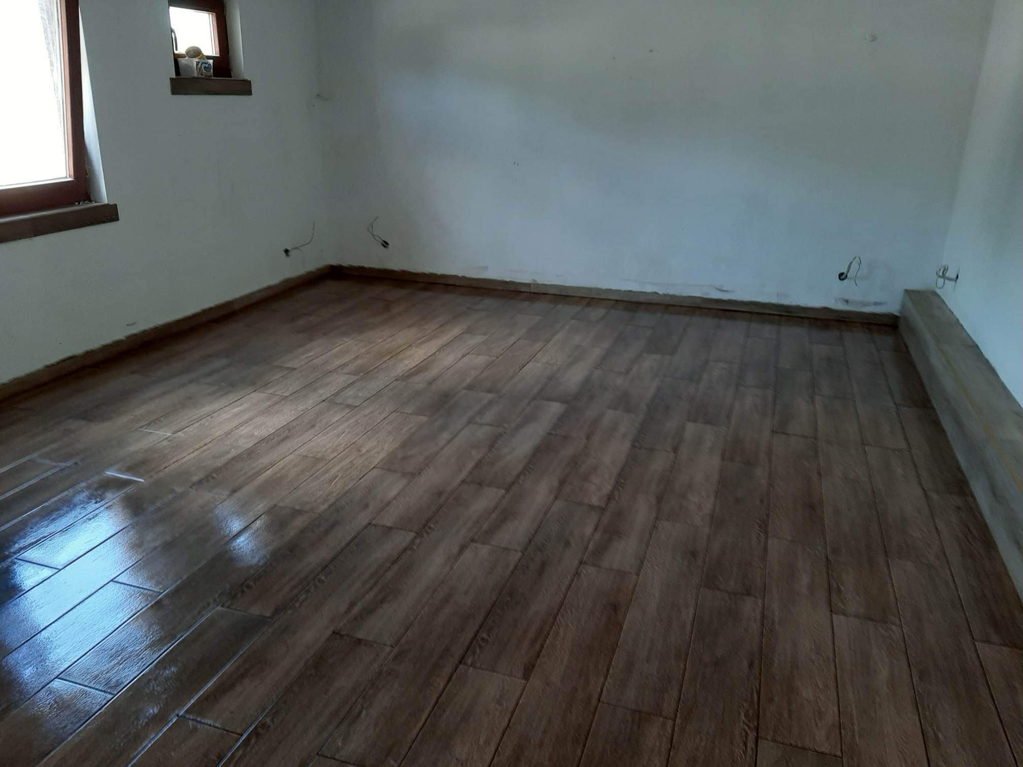 Ułożona, brązowa podłoga z płytek imitujących drewno w pustym pomieszczeniu z białymi ścianami. Widoczne niedociągnięcia przy ścianach i wystające kable.