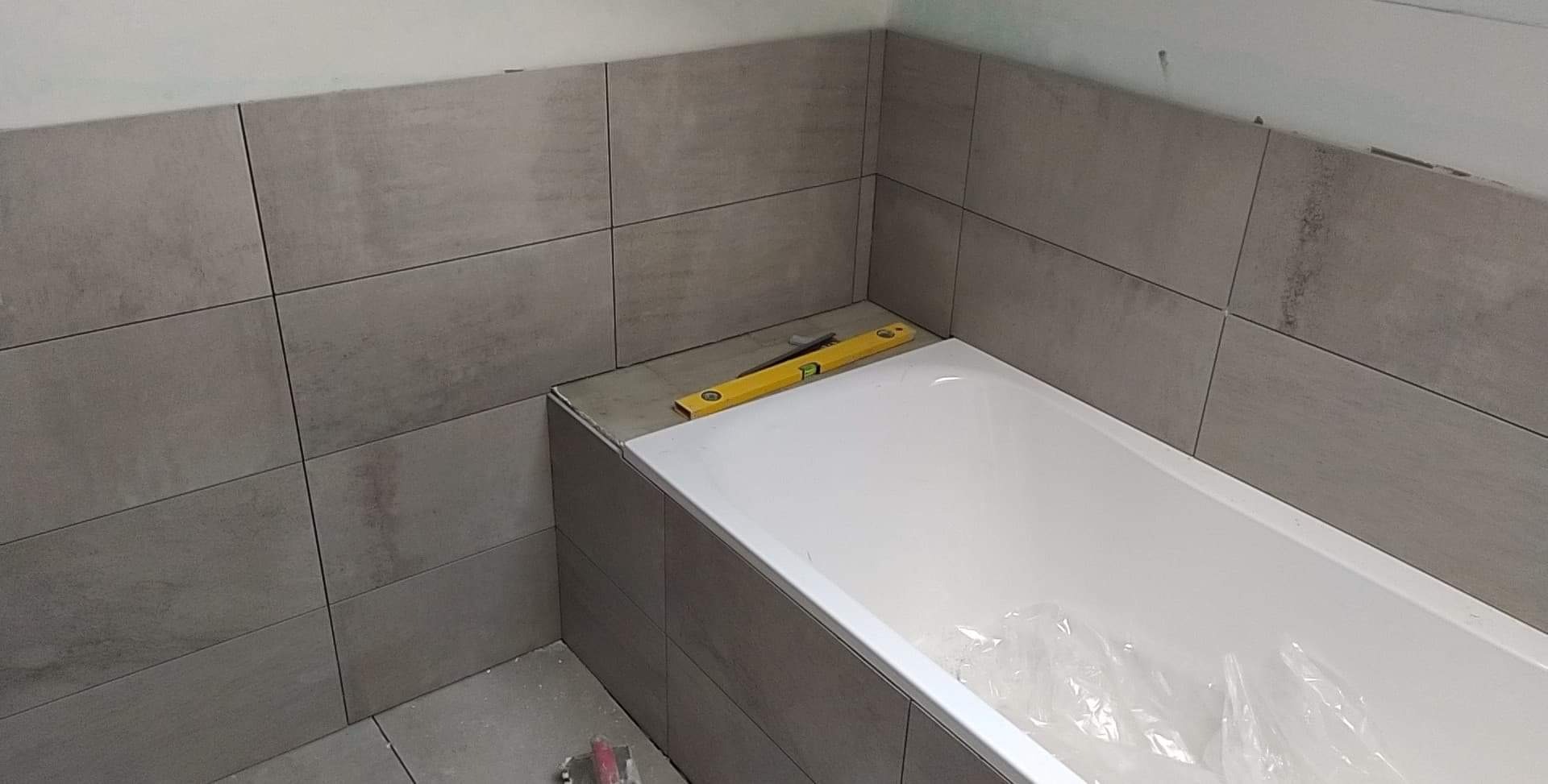 Szare, prostokątne płytki ułożone wokół wanny, z żółtą poziomicą leżącą na krawędzi obudowy wanny, częściowo przykrytej folią ochronną.