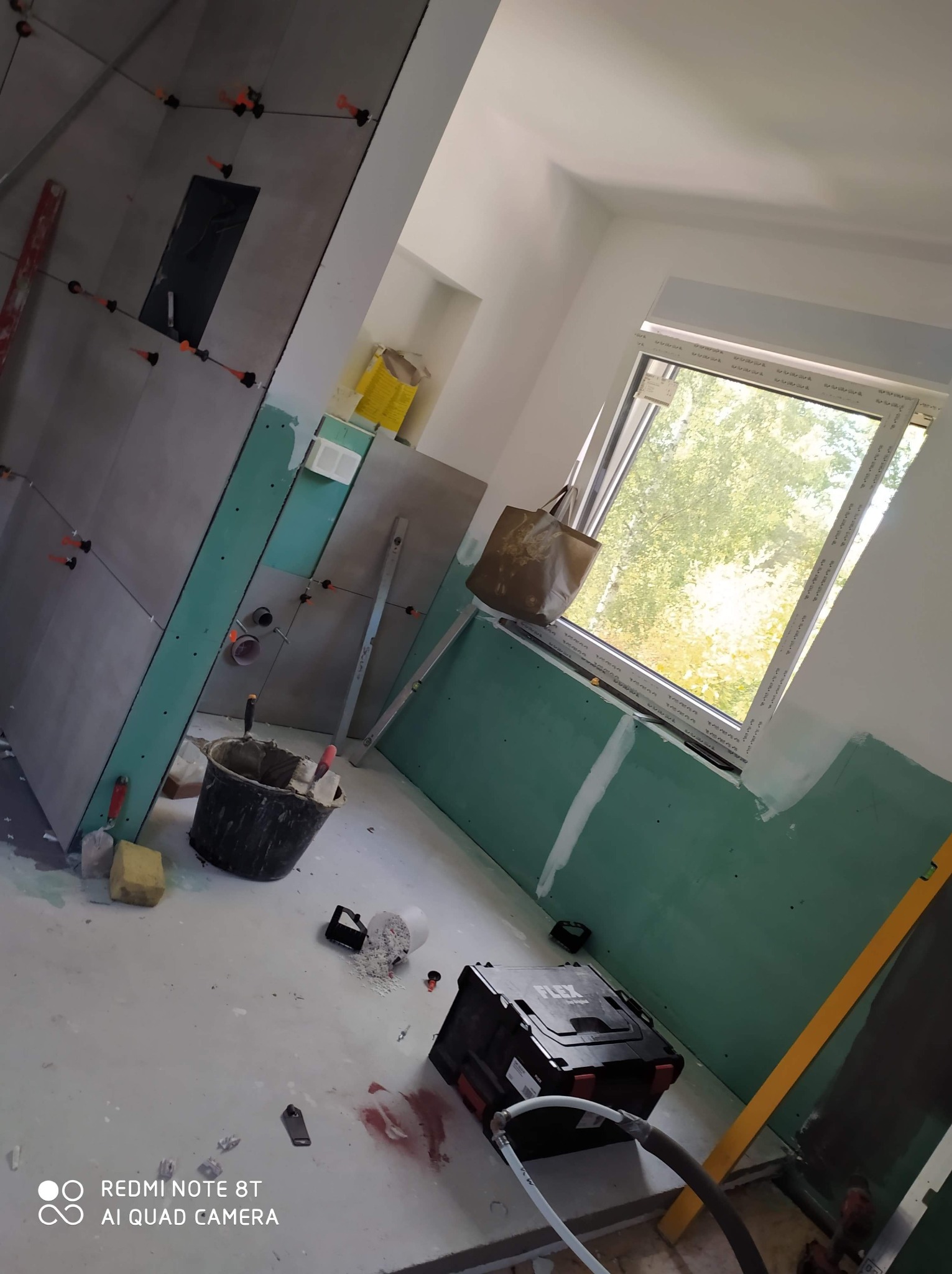 Wnętrze łazienki w trakcie remontu: szare płytki częściowo ułożone na ścianie z otworem, płyty kartonowo-gipsowe na ścianach, narzędzia i wiadro na podłodze, widok na zieleń za oknem.