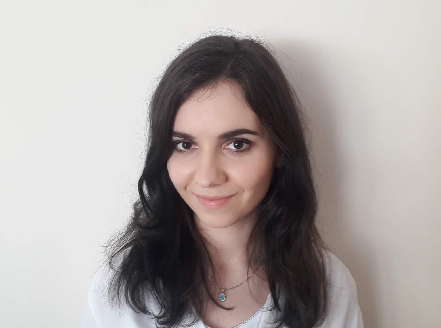 Portret uśmiechniętej młodej kobiety z ciemnymi włosami i delikatnym makijażem, prezentującej się jako specjalistka z Lubaczowa, na tle jasnej ściany, z widocznym subtelnym naszyjnikiem...