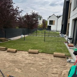 Patryk Parzys - Układanie trawy z rolki w ogrodzie nowoczesnego domu. Częściowo rozłożona darń, widoczne narzędzia i rolki trawy. Czarny płot w tle, pochmurne niebo.