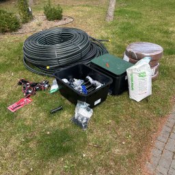 Patryk Parzys - Elementy systemu nawadniania rozłożone na trawie: rura w zwoju, złączki, skrzynka sterująca i nawóz. Przygotowanie do instalacji systemu nawadniania ogrodu.
