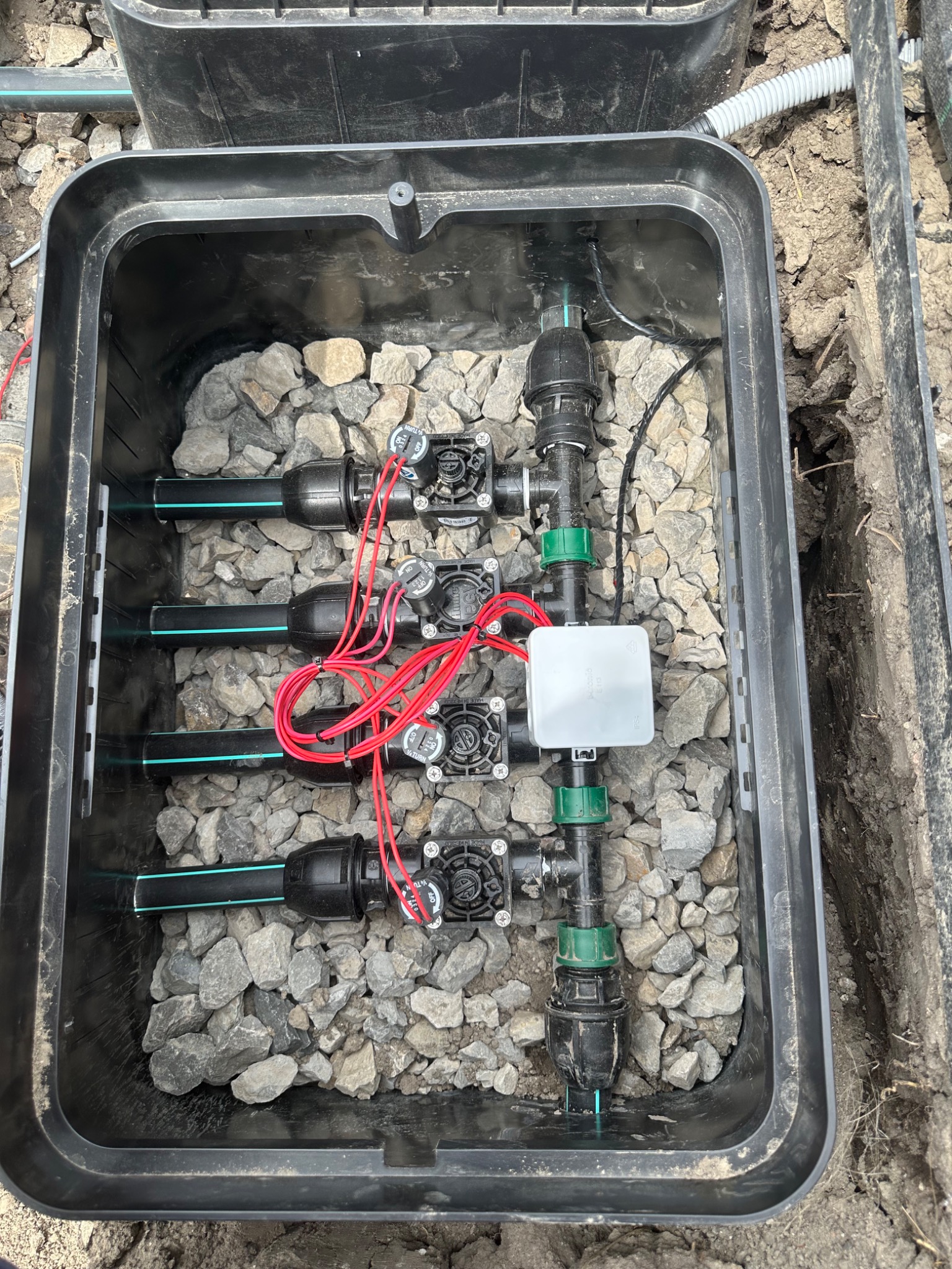 Skrzynka zaworowa z systemem nawadniania, wypełniona kamieniami. Widoczne rury, zawory i przewody elektryczne. Instalacja w czarnej skrzynce, ziemia dookoła.
